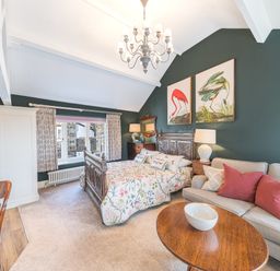 A bedroom with a wooden bed, floral bedding, a beige sofa with red and beige pillows, wooden tables, and bird paintings on a green wall at Prospect Cottage Loft in Ambleside