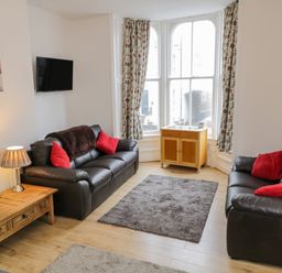 A living room with two black sofas with red cushions a wooden coffee table a small wooden cabinet under a large window and a mounted flat screen TV at 1 Church Square Whitby