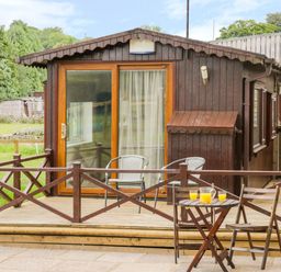 A wooden cabin with a deck and sliding glass door with two chairs on the deck and a small table with two chairs and a jug of orange juice in front at Thirley Beck Lodge in Harwood Dale