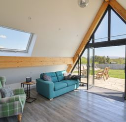 A living room with a green plaid armchair a blue sofa and large triangular windows opening to a patio at Elan Valley Welsh - The Sheepfold in Llanyre near Llandrindod Wells
