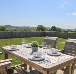 An outdoor wooden dining table set with plates bowls cutlery wine glasses and a plant with a fenced grass yard and hills in the background at Elan Valley Welsh - The Sheepfold in Llanyre near Llandrindod Wells