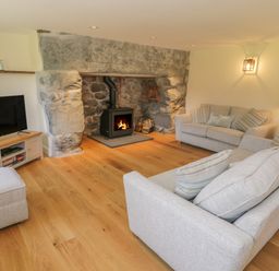 A living room with a stone fireplace wood stove two gray sofas a TV on a wooden stand and hardwood flooring at Tyn Ffynnon in Dyffryn Ardudwy