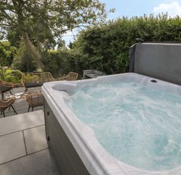 An outdoor patio area with a hot tub and seating with chairs and a bench surrounded by greenery at Argraig in Llanbedrog