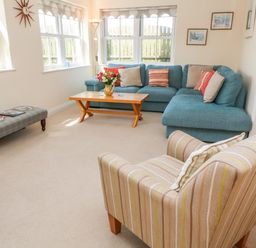A living room with a blue corner sofa, a wooden coffee table, an upholstered armchair, a bench, and windows at Henrys Croft in Lizard
