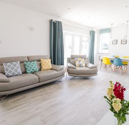 A living room with beige sofas and colorful cushions next to a dining table with multicolored chairs and flowers on a table at Senafe in Widemouth Bay