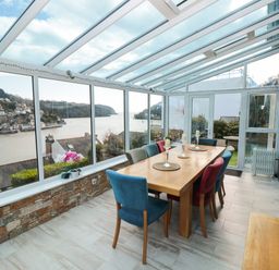 A dining area with a wooden table and colorful chairs in a glass conservatory overlooking a river and houses at Estuary View House and Annexe in Dartmouth