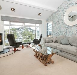 A living room with a gray sofa a glass coffee table with a wooden base two black chairs near large windows and a wall-mounted round mirror at Estuary View House and Annexe in Dartmouth
