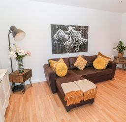 A living room with a brown sofa with yellow cushions and a blanket a floor lamp a side table with flowers a wall clock and a plant at The Snug in Beadnell