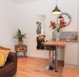 A small dining area with a wooden table, two black bar stools, a vase with flowers, a brown sofa with yellow cushions, a plant, and a wavy mirror on the wall at The Snug in Beadnell