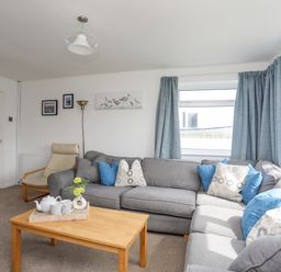A living room with a gray sectional sofa, blue and patterned cushions, a wooden coffee table with a tray, a beige armchair, framed pictures, and blue curtains at Crows Nest - Rhosneigr in Rhosneigr