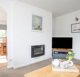 A living room with a wall-mounted electric fireplace a television on a stand wooden table with a tray teapot cups and a vase with flowers and a dining area with wooden chairs and table through an archway at Crows Nest - Rhosneigr in Rhosneigr