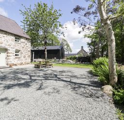 A stone building with a light door and windows next to a gravel area with a wooden picnic table surrounded by trees and plants at Stabla in Chwilog