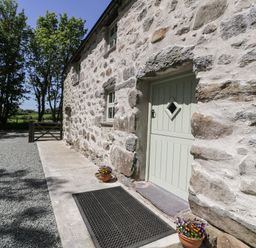 Stone exterior wall of a building with a green door and small windows next to a gravel pathway with flower pots at Stabla in Chwilog