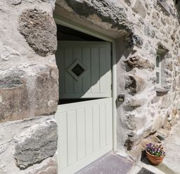 A half open pale green stable door set in a rustic stone wall with a diamond shaped window and a flower pot on the ground at Stabla in Chwilog