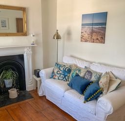 A white couch with decorative pillows in front of a wall with beach artwork beside a white fireplace with a plant inside at The Ferryman in Weymouth