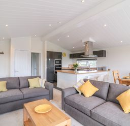 A living room with gray sofas and yellow pillows next to a kitchen with wooden counters and a dining table at Alder Lodge in Teigngrace near Newton Abbot