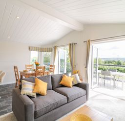 A living room with a gray sofa and yellow pillows next to a dining area with wooden chairs and table at Alder Lodge in Teigngrace near Newton Abbot