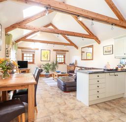 An open plan living room with wooden dining table, leather chairs, kitchen island, washing machine, and wooden beams on the ceiling at Valley View in St Martin near Mawgan