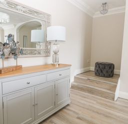 A hallway with a grey sideboard with a lamp and decorative metal art and a mirror above it with wooden steps and a grey cushion at Harbour Watch 2 in Weymouth