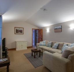 A living room with a beige sofa and blue and white pillows a small wooden coffee table on a rug a television on a stand a wooden side table with magazines curtains on a window and two framed pictures on walls at Riverhouse Lodge in Upwey