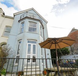 The rear exterior of a light blue house with upper balcony and patio table with umbrella at Elmbank in Looe