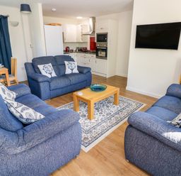 A living room with blue sofas a wooden coffee table a wall-mounted television and a kitchen area at Little Freestone in Carew