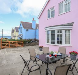 Outdoor patio area with table and chairs beside a pink house near a seaside road at The Pink House in Rhosneigr