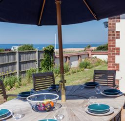 An outdoor dining table with six place settings under a large umbrella near a garden with a view of the sea at Beach Haven in West Bexington