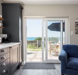 A kitchen area with dark cabinets and a coffee machine next to a sliding glass door opening to a patio with wooden chairs and an umbrella at Beach Haven in West Bexington