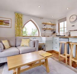 A living room with a gray sofa yellow pillows wooden coffee table and a kitchen with wooden stools and white cabinets at The Folly in Weobley