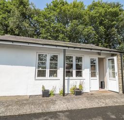 A small white building with three windows and two open doors with plants outside and trees in the background at Vallum Kiosk in Haltwhistle