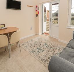 A small living room with a dining table and two chairs a wall-mounted tv a gray sofa a patterned rug and a door leading outside at Vallum Kiosk in Haltwhistle