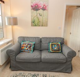 A gray sofa with two colorful cushions under a floral painting beside a window with a floor lamp at Vallum Kiosk in Haltwhistle