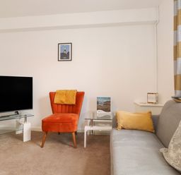 A living room with a grey sofa and orange chair with a glass TV stand and framed picture on the wall at Holly Cottage in Keswick