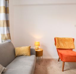 A living room with a gray sofa with cushions a small round side table with a lamp and an orange armchair with a blanket at Holly Cottage in Keswick