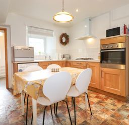 A kitchen with a table covered by a yellow tablecloth surrounded by white chairs and wooden cabinets with appliances at Holly Cottage in Keswick