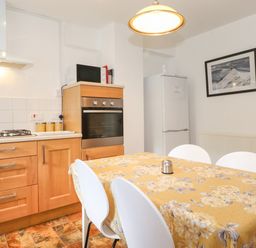 A kitchen with wooden cabinets a gas stove an oven a microwave and a refrigerator with a dining table covered by a floral tablecloth and white chairs at Holly Cottage in Keswick
