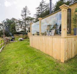 A wooden cabin with a deck and glass railing on a green lawn with outdoor barbecue and trees at High Rigg Shepherd's Retreat in Brampton Cumbria