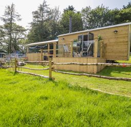 A wooden cabin with a small deck and outdoor chairs surrounded by a rustic wooden fence and green grass at High Rigg Shepherd's Retreat in Brampton Cumbria