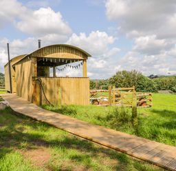 A wooden shepherds hut with a curved roof and a wooden walkway leading to it in a grassy field with a wooden gate at High Rigg Shepherd's Retreat in Brampton Cumbria