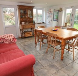 A kitchen dining area with a wooden table and chairs a red sofa a wooden cabinet and a refrigerator with windows and doors showing a garden at Kernyk in Rinsey near Porthleven