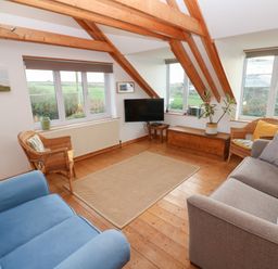 A living room with wooden beams a blue armchair two wicker chairs a gray sofa and a wall-mounted TV at Kernyk in Rinsey near Porthleven