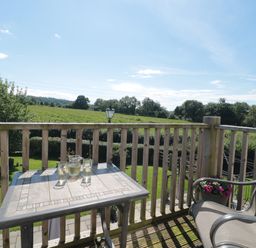 A balcony with a table and chairs overlooking a green field with trees and a blue sky at The Cider Shed in Winscombe