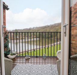 A balcony with a patterned rug and a wicker chair overlooking a river and trees at Maplebeck in Whitby