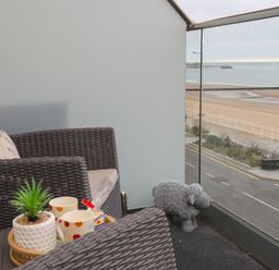 A balcony with two woven chairs a tray with cups a plant and a sheep figurine overlooking a beach at Puffin View in Rhos On Sea