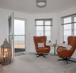 A seating area with two brown armchairs and a round glass table near windows overlooking a beach at Puffin View in Rhos-On-Sea