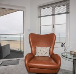 A brown armchair with a cushion next to a glass side table and open door leading to a balcony with a view of the beach and sea at Puffin View in Rhos-On-Sea