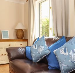 A living room with a brown leather sofa and blue cushions next to a window and a white sideboard with a lamp and flowers at Deja Blue in Falmouth