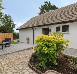 An outdoor patio area with a wooden deck and black patio furniture next to a white house with a brown tiled roof and garden bed at Little Valley View in Angarrack