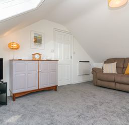 A carpeted room with a brown sofa with two cushions a white door a cabinet with a lamp clock and glass dish and a TV on a black stand at Little Valley View in Angarrack